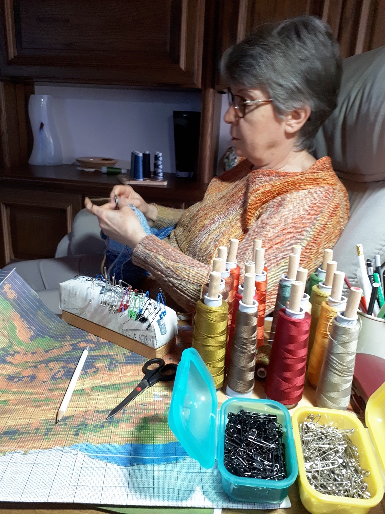 Gabriele Tippel am Arbeitsplatz-KlugeStrickArt Gabriele Tippel gemütlich im Sessel sitzend und an einem Wandbild, einer gestrickten Landschaft in KlugeStrickArt arbeitend